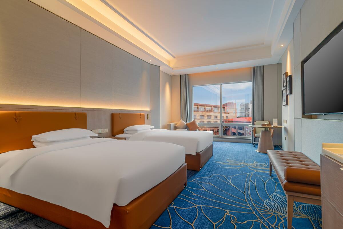 Habitación de hotel con dos camas, un escritorio, una silla y un ventanal con vistas a la ciudad.