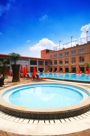 Zona de piscina de hotel con una piscina grande, tumbonas y sombrillas rojas.