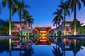 Entrada de hotel con una piscina reflectante, palmeras y una fachada bien iluminada.