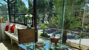 A glass-top table with wooden legs, a bench with red cushions, and a view of a garden through a glass window.