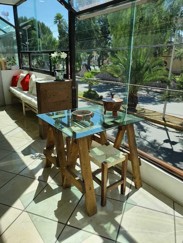 Une table dont le plateau est en verre et les pieds en bois, un banc avec des coussins rouges, et une vue sur un jardin à travers une fenêtre en verre.
