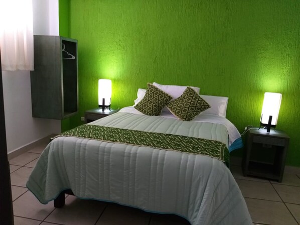 A bed with a white quilt and green patterned pillows, two bedside tables with lamps, a green wall, and a wardrobe.