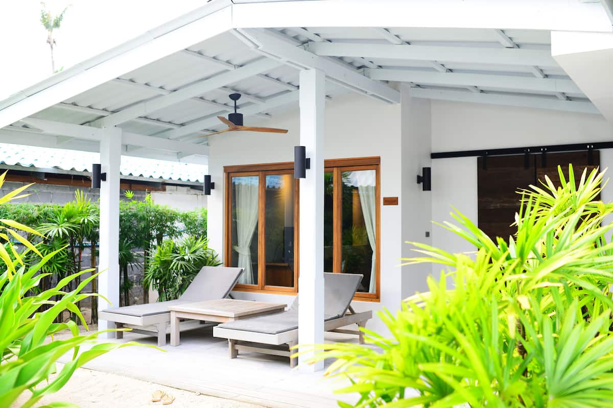 A covered outdoor seating area with lounge chairs and a ceiling fan, surrounded by lush greenery.