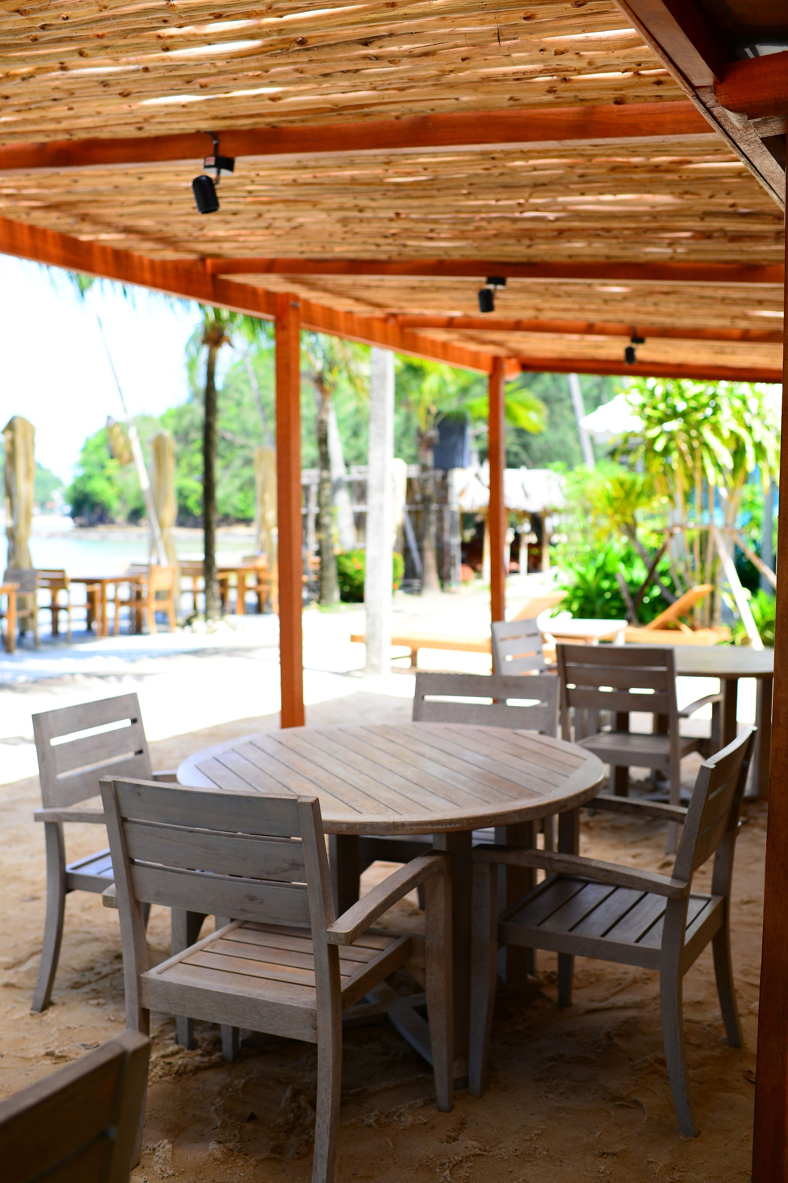 A covered outdoor seating area with wooden tables and chairs, overlooking a beach and palm trees.