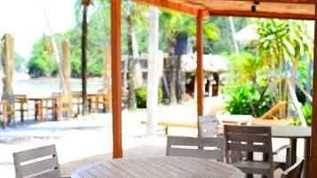 A covered outdoor seating area with wooden tables and chairs, overlooking a beach and palm trees.