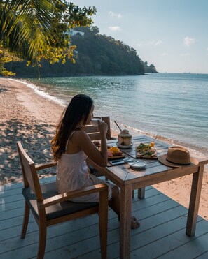 A woman sitting at a wooden table on a deck by the sea, with a meal and a straw hat on the table. - The Cove Phuket  (Wichit)