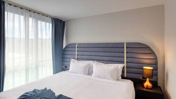 A hotel room with a large bed, a headboard with a geometric pattern, a bedside lamp, and sheer curtains on the window.