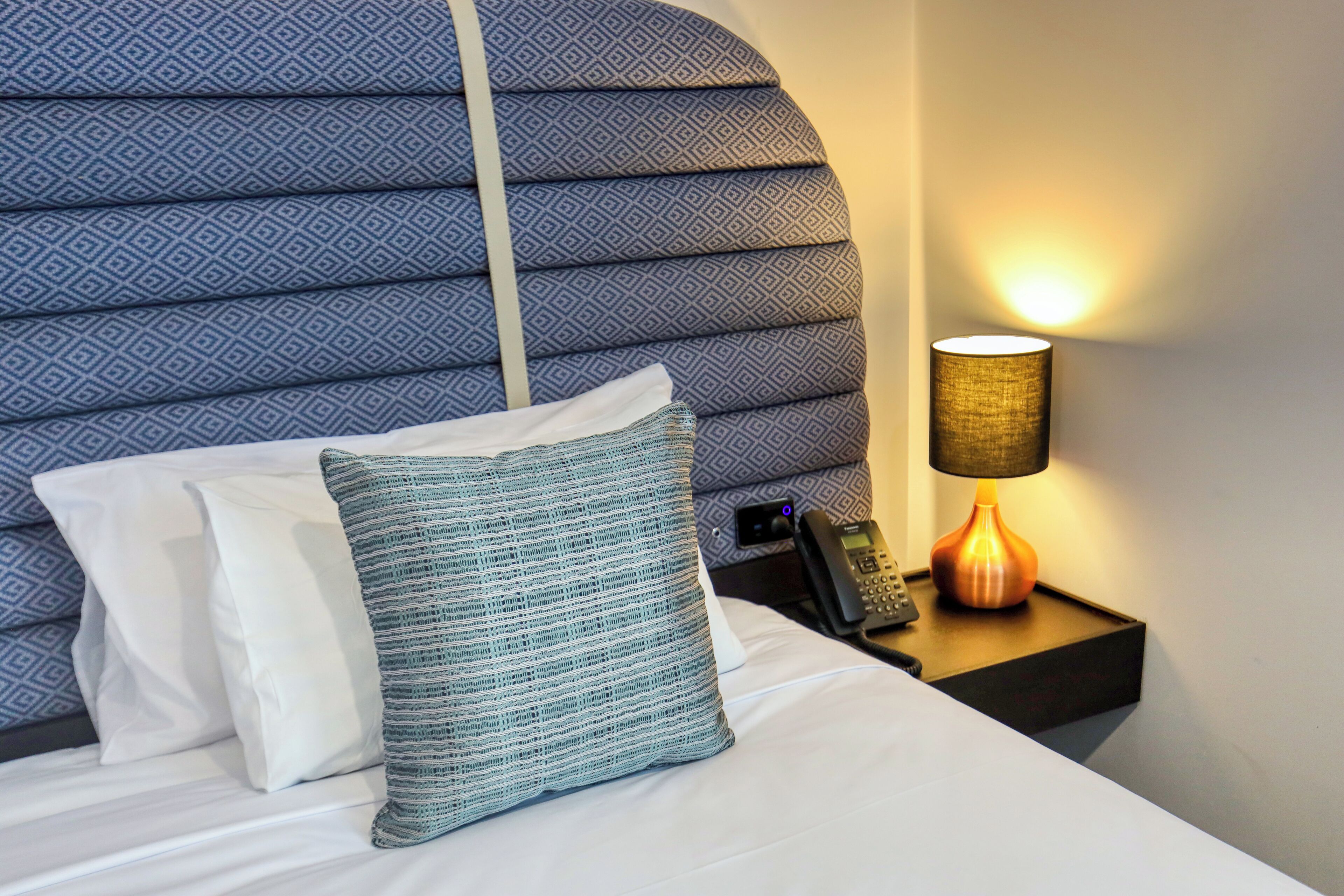 A neatly made bed with white and blue pillows, a patterned headboard, a bedside lamp, and a phone on a nightstand.