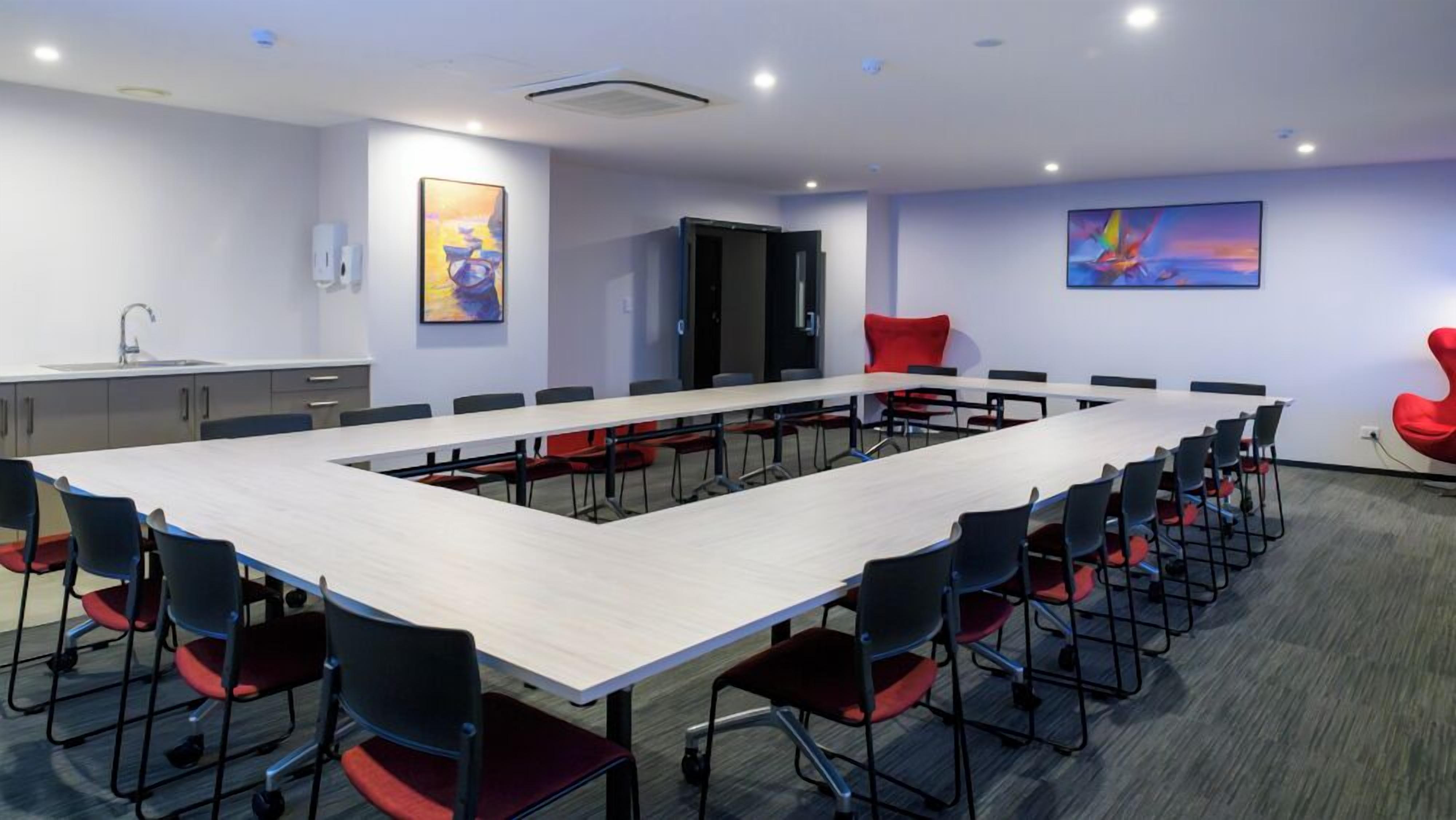 A conference room with a long table, black chairs with red cushions, a sink, cabinets, and two paintings on the walls.