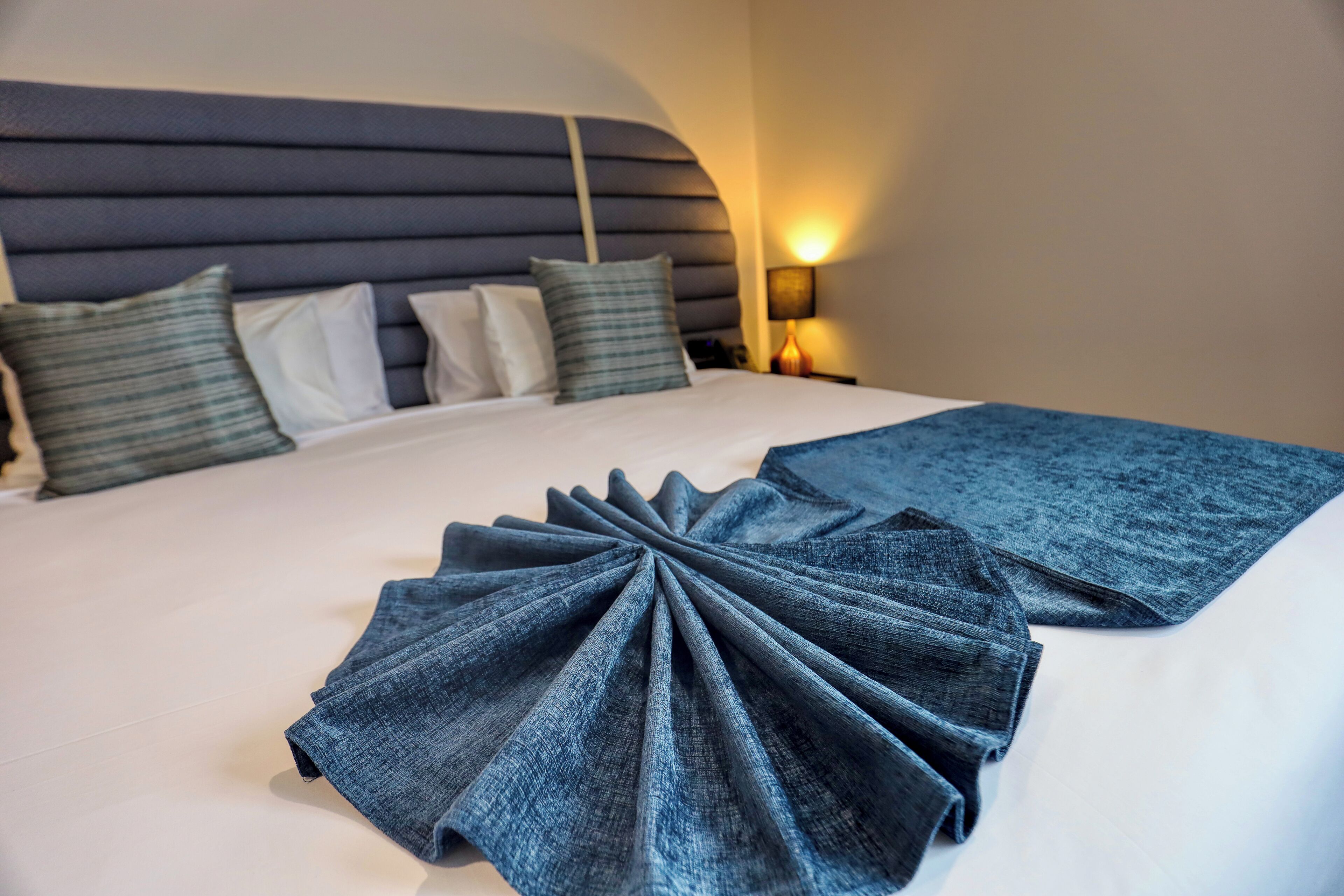 A neatly made bed with a blue blanket and pillows, a bedside lamp, and a headboard.