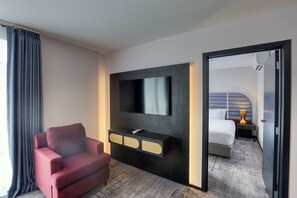 A hotel room with a flat-screen TV mounted on a dark wall, a red armchair, a bedside table with a lamp, and a bed with a headboard. - Marsden Suites Albany (Auckland)