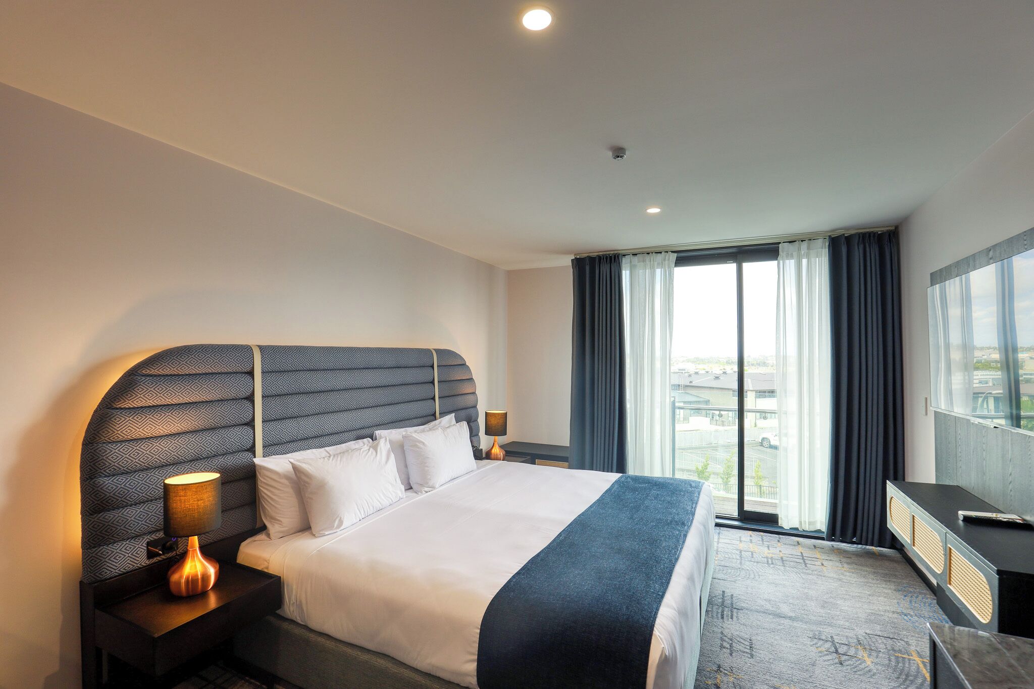 A hotel room with a large bed, a nightstand with a lamp, a TV, and a view of the city through the window.