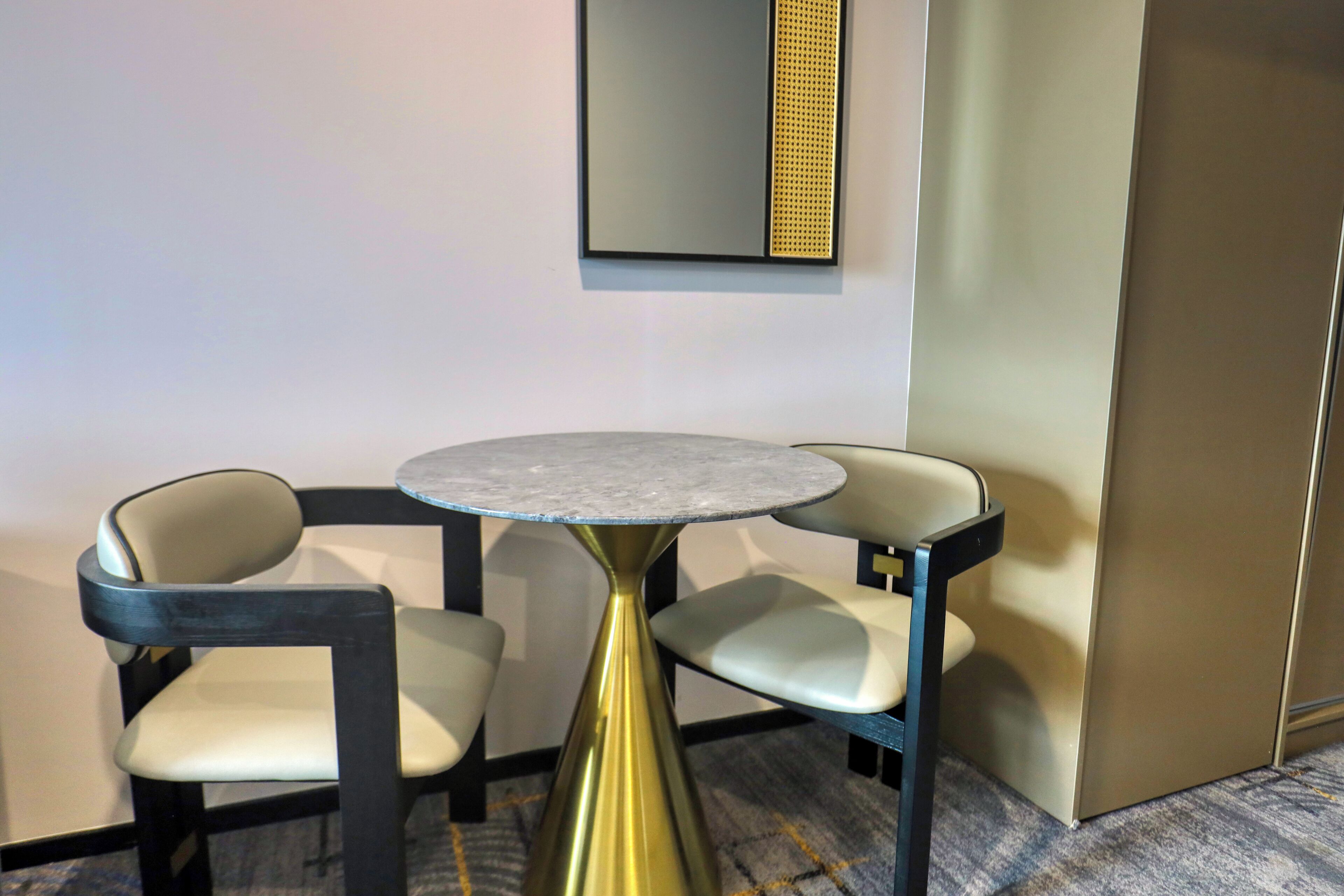 A small, round table with a metallic base, two chairs with cushioned seats, and a wall-mounted mirror with a decorative panel.