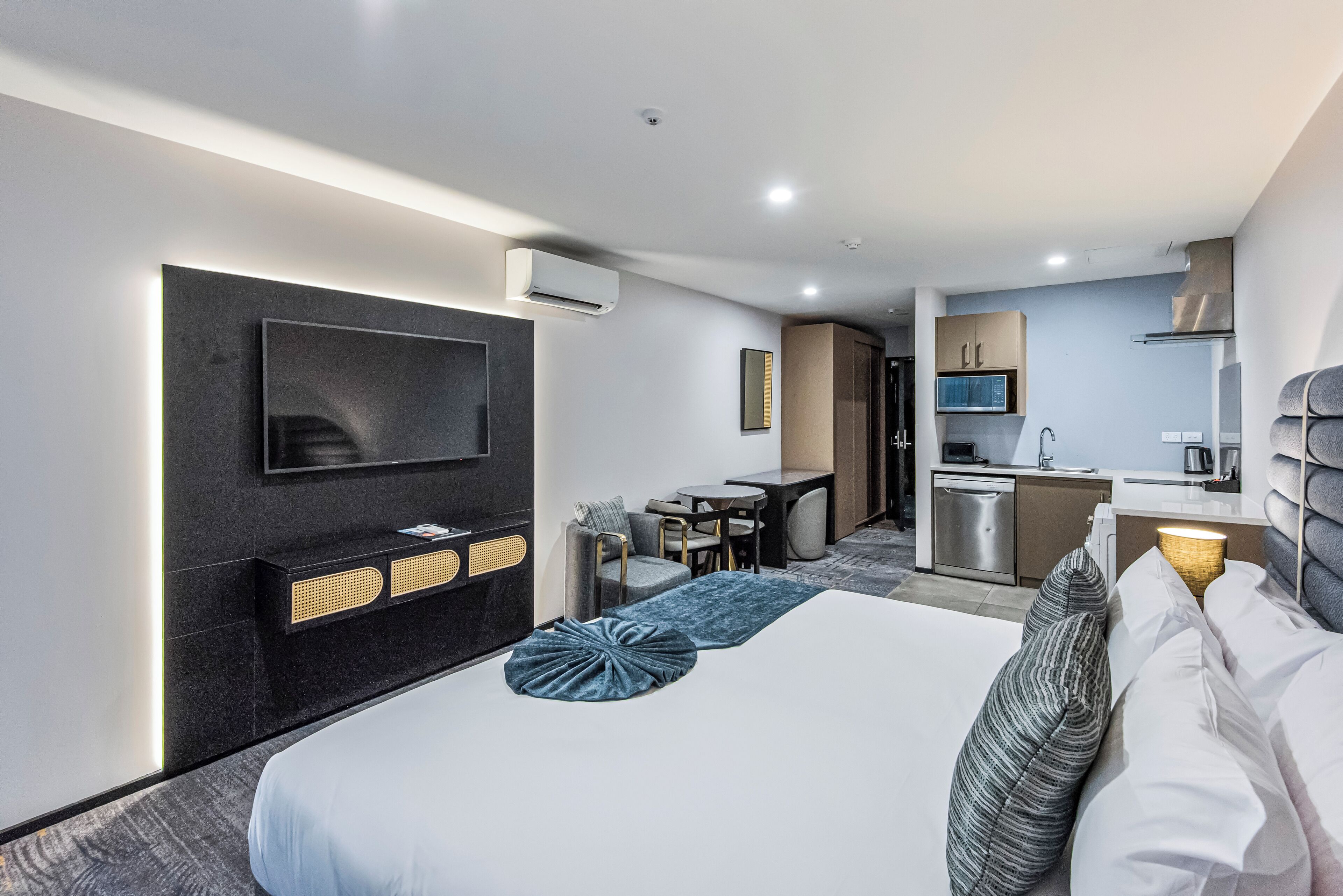 A modern hotel room with a large bed, a flat-screen TV mounted on the wall, a small kitchenette, and a seating area with a table and chairs.