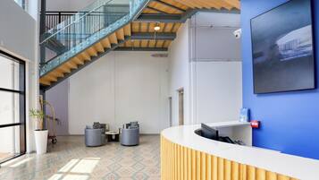 Un área de recepción moderna con un mostrador de madera, una pared de acento azul, una escalera con barandillas de vidrio y un ventanal que deja entrar luz natural.
