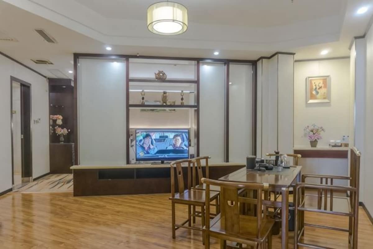 Un área de comedor con una mesa y sillas de madera, un televisor sobre un mueble bajo y un estante decorativo con objetos.
