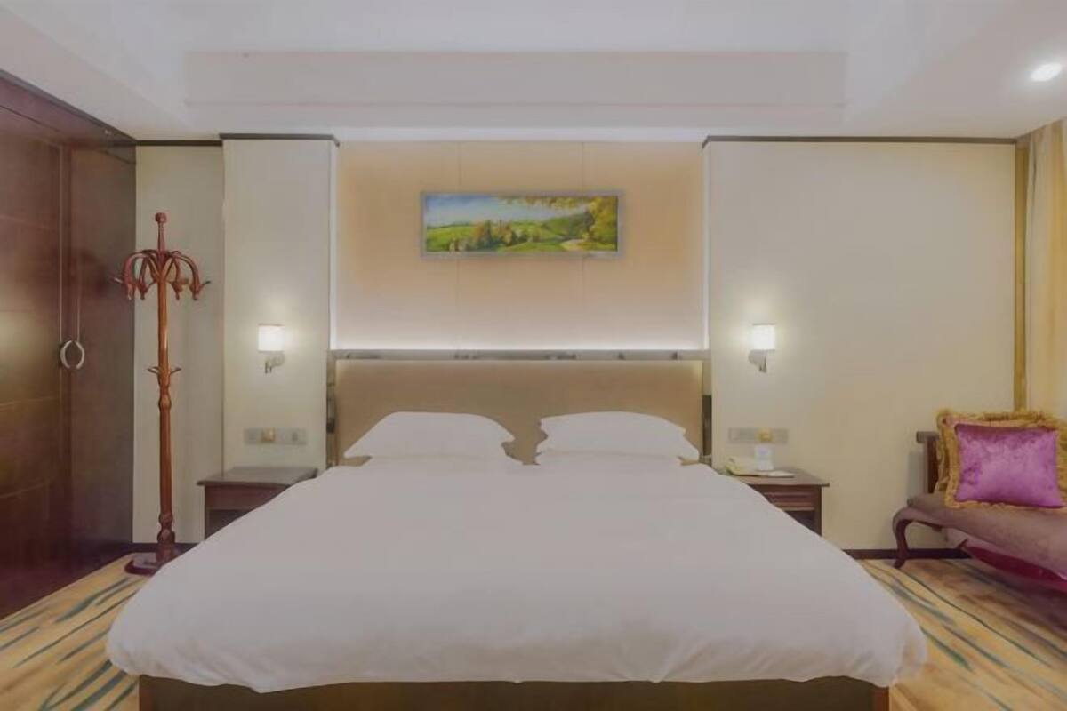 Une chambre d’hôtel avec un grand lit, deux tables de chevet, une commode, une lampe, un tableau au mur et une chaise.