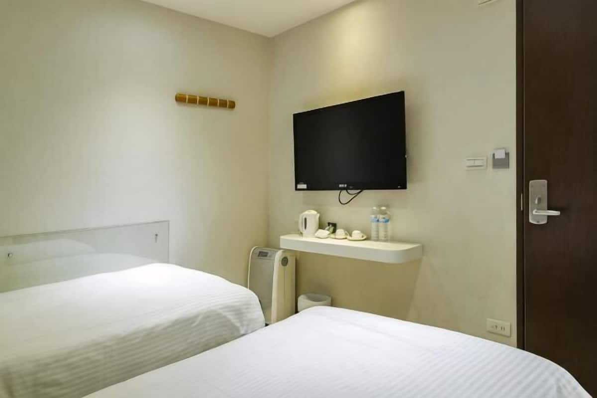 A hotel room with two beds, a TV mounted on the wall, a small shelf with toiletries, and a door.
