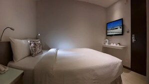 Habitación de hotel con cama, mesita de noche con lámpara, televisor montado en la pared y estante pequeño con artículos de aseo.