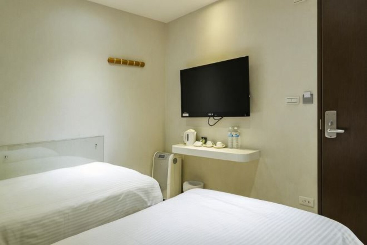 A hotel room with two beds, a TV mounted on the wall, a small shelf with toiletries, and a door.