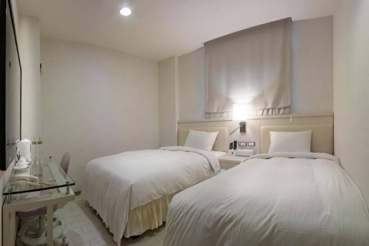 A hotel room with two beds, a desk with a chair, a bedside table, and a wall-mounted lamp.