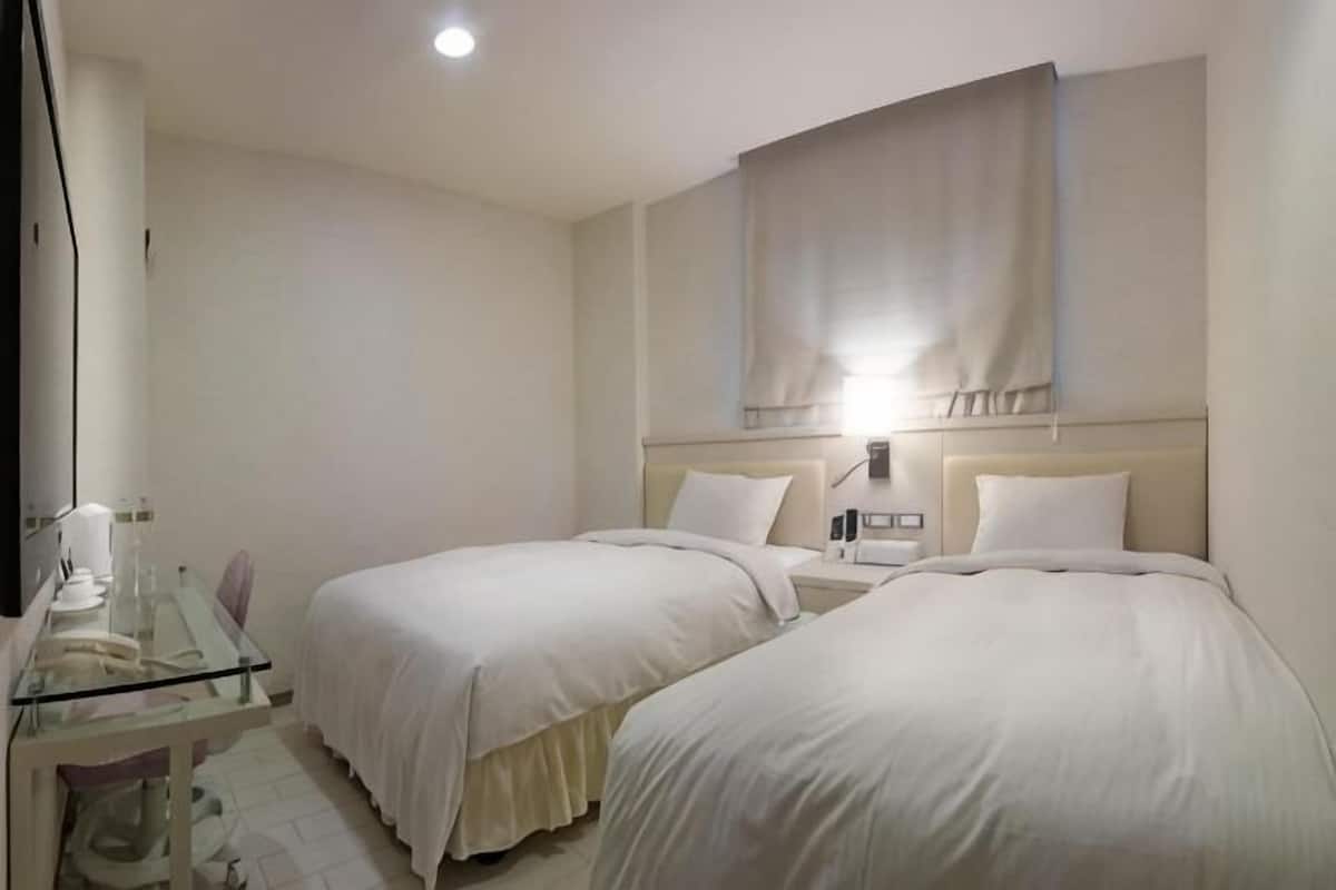A hotel room with two beds, a desk with a chair, a bedside table, and a wall-mounted lamp.