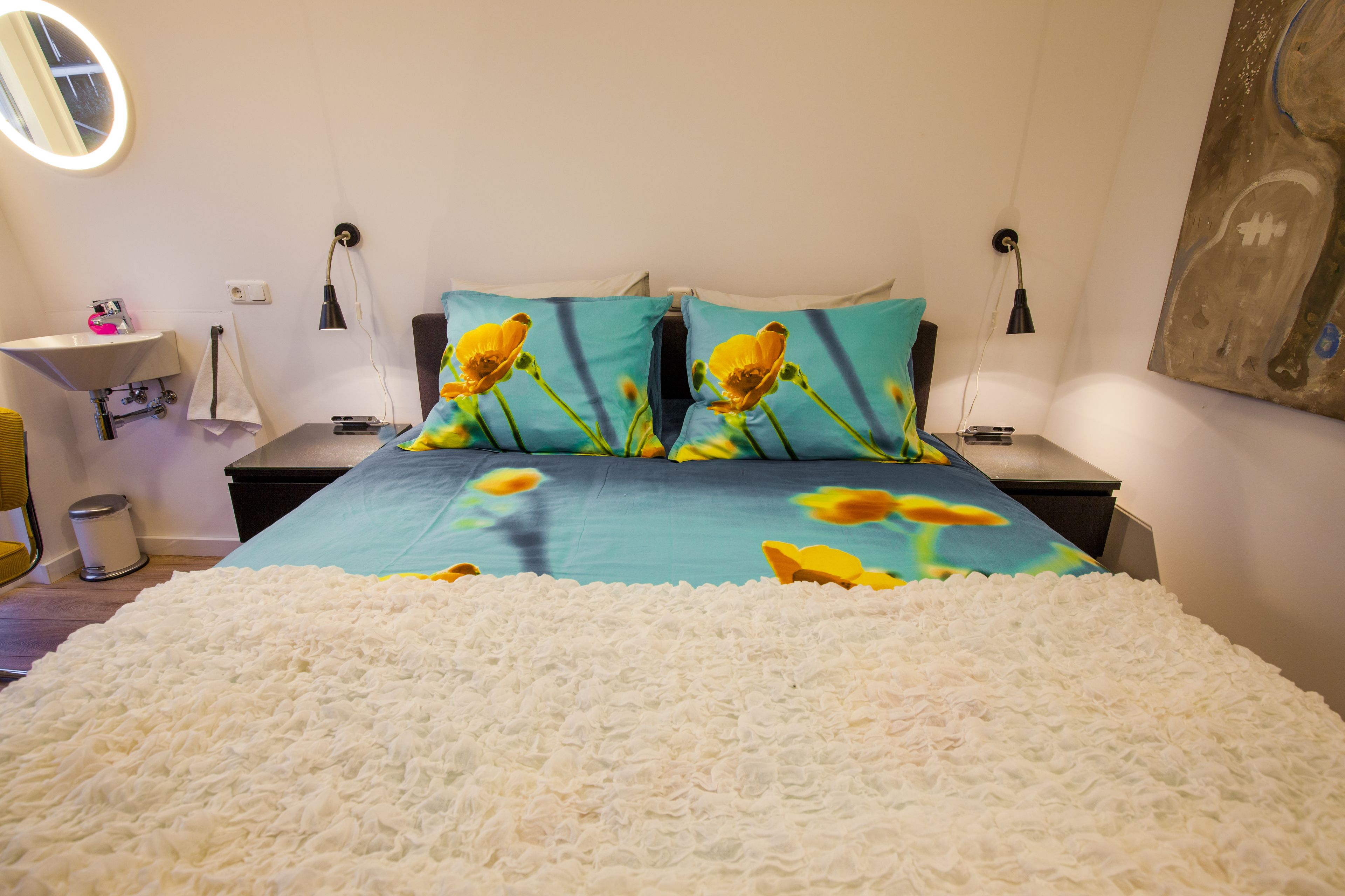 Une chambre à coucher comprenant un lit, deux tables de chevet, une commode, un miroir, une lampe fixée au mur et un tableau.
