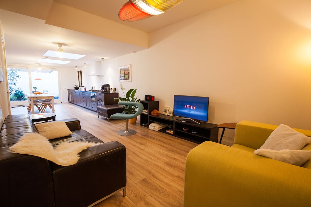 Una sala moderna con un sofá, un sillón amarillo, un televisor y un comedor con mesa y sillas.