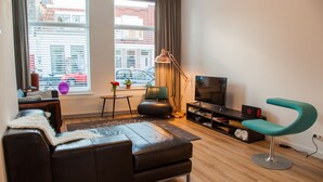 Een moderne woonkamer met een leren bank, een turquoise fauteuil, een vloerlamp, een televisie en uitzicht op de stad door grote ramen.