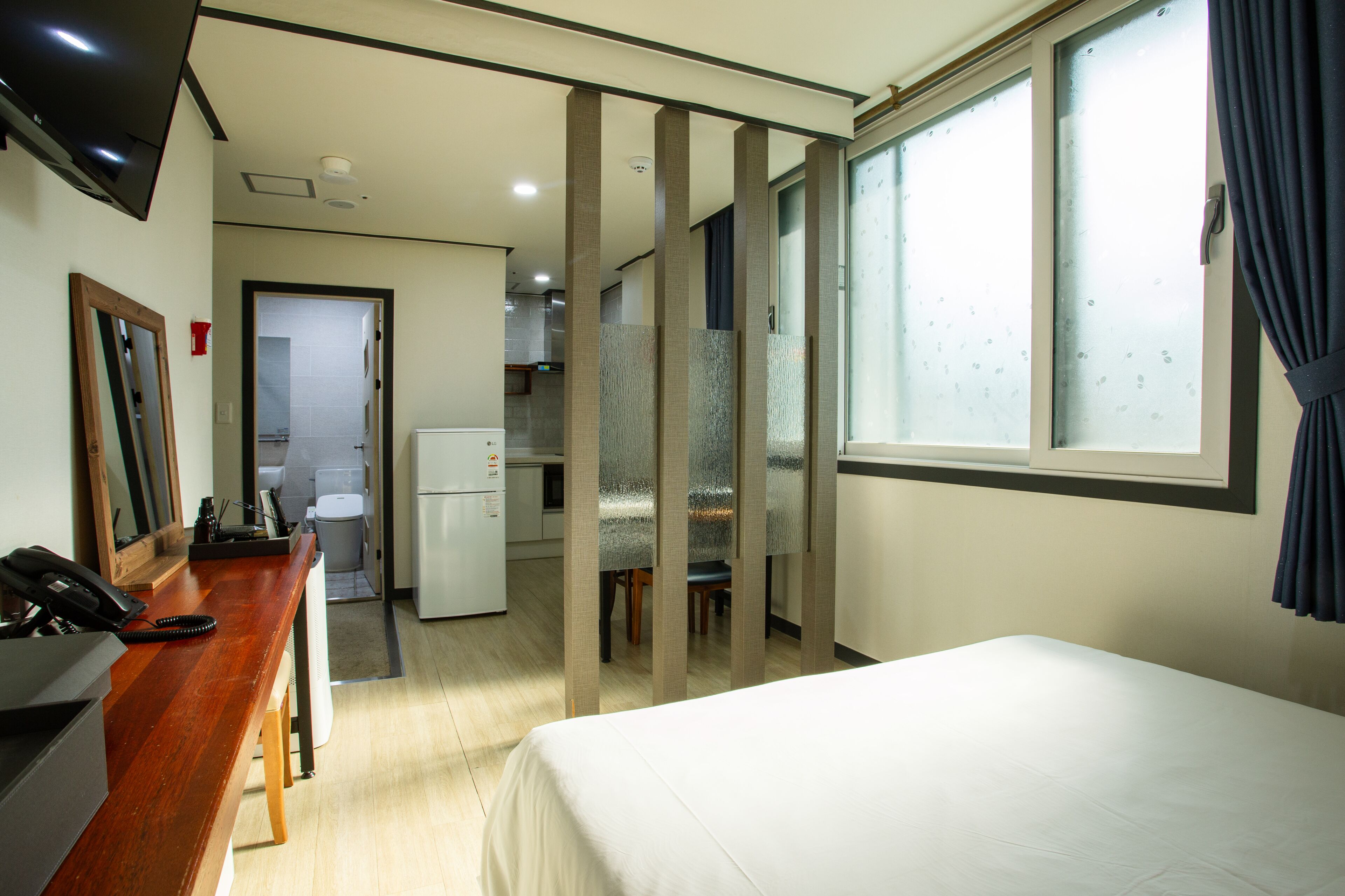 Habitación de hotel con cama, escritorio con teléfono, espejo, televisor montado en la pared y vista a la cocina y el baño a través de puertas con vidrio esmerilado.