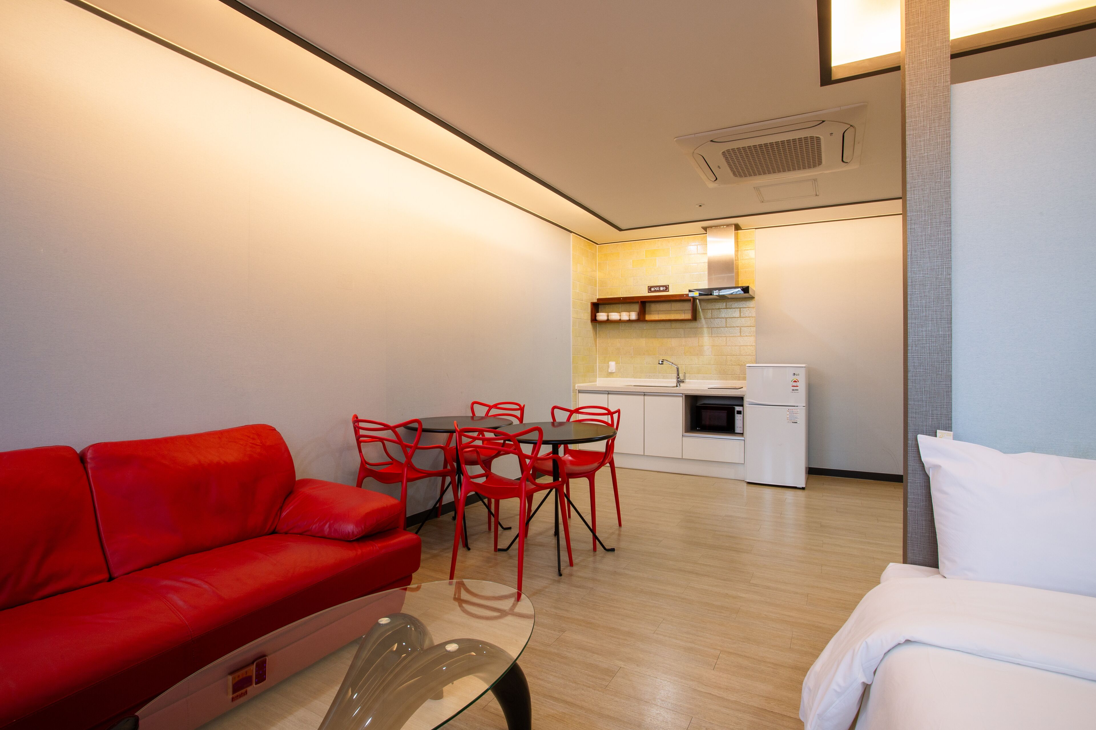 A compact living space with a red sofa, a glass coffee table, a white bed, and a small kitchenette in the background.
