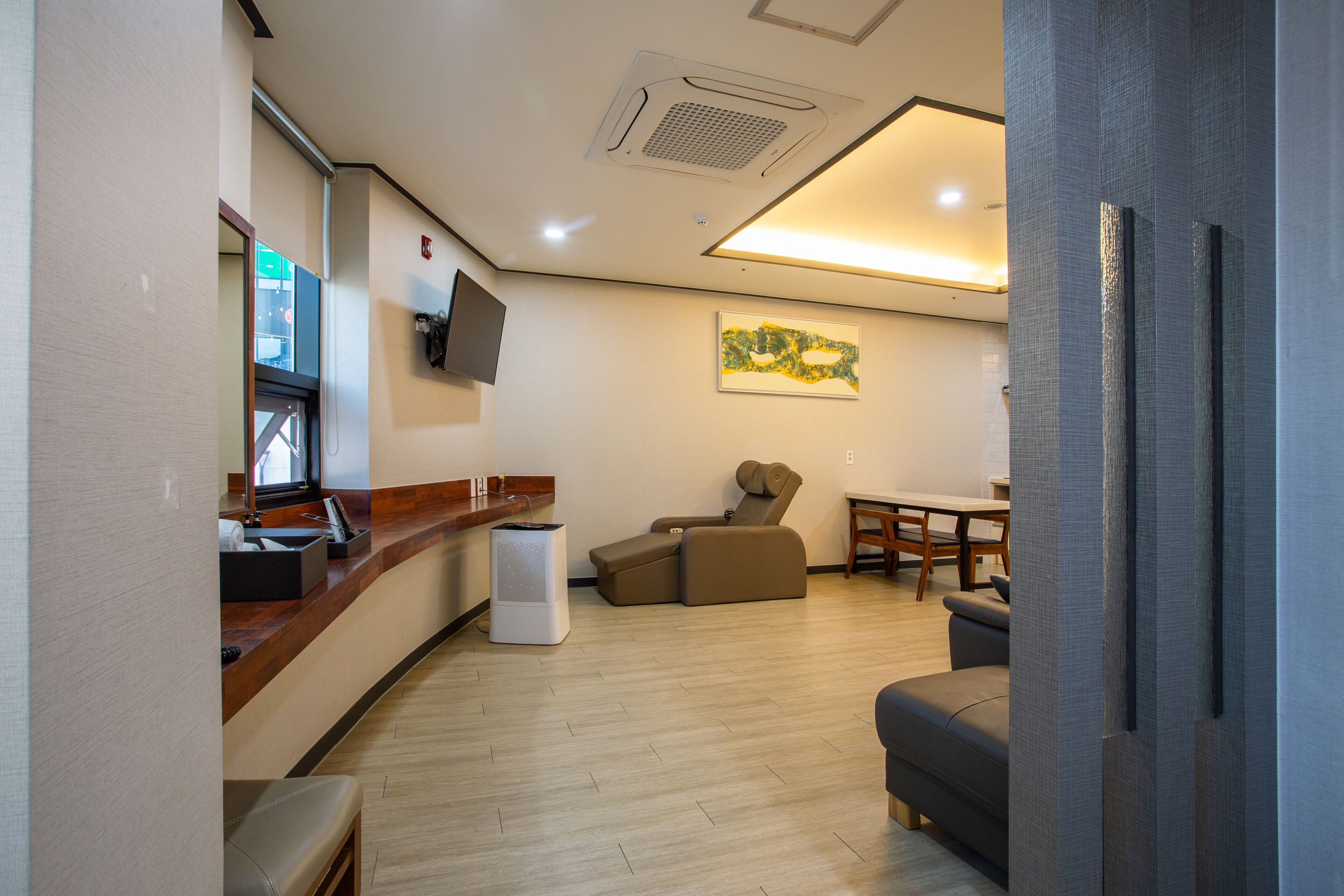 A modern hotel room with a wooden reception desk, a seating area with a sofa and armchairs, a dining table, and a large painting on the wall.