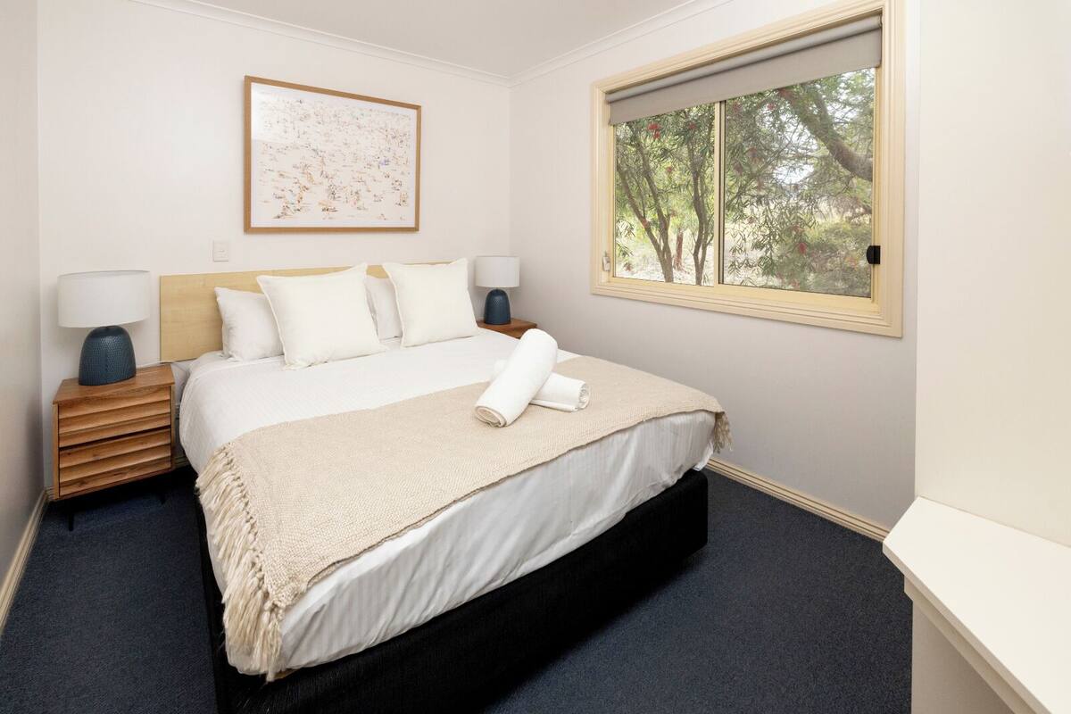 Een opgemaakt bed met witte lakens en een beige plaid, een houten nachtkastje met een lamp, een lijst met een kunstwerk aan de muur en een raam met uitzicht op bomen.