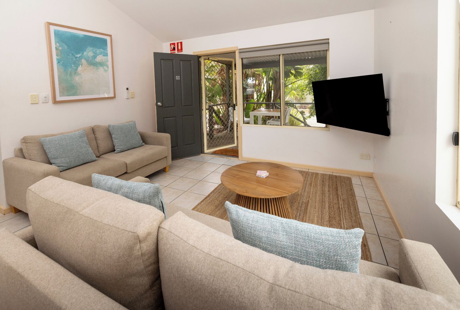 A living room with a sofa set, a coffee table, a TV mounted on the wall, and a sliding glass door leading to a balcony.