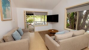 Un salon avec deux canapés beige, une table basse, un téléviseur à écran plat fixé au mur et une porte-fenêtre coulissante donnant sur un espace extérieur.