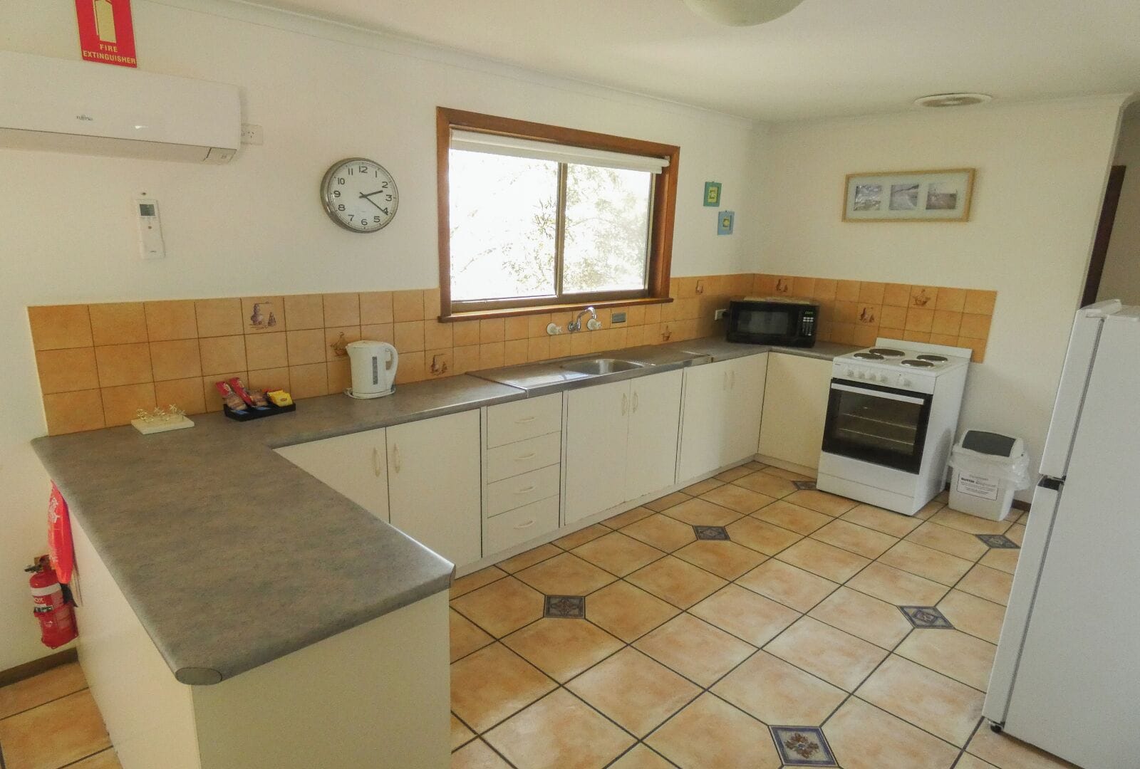 A kitchen with a microwave, stove, and sink, a clock on the wall, and a window.