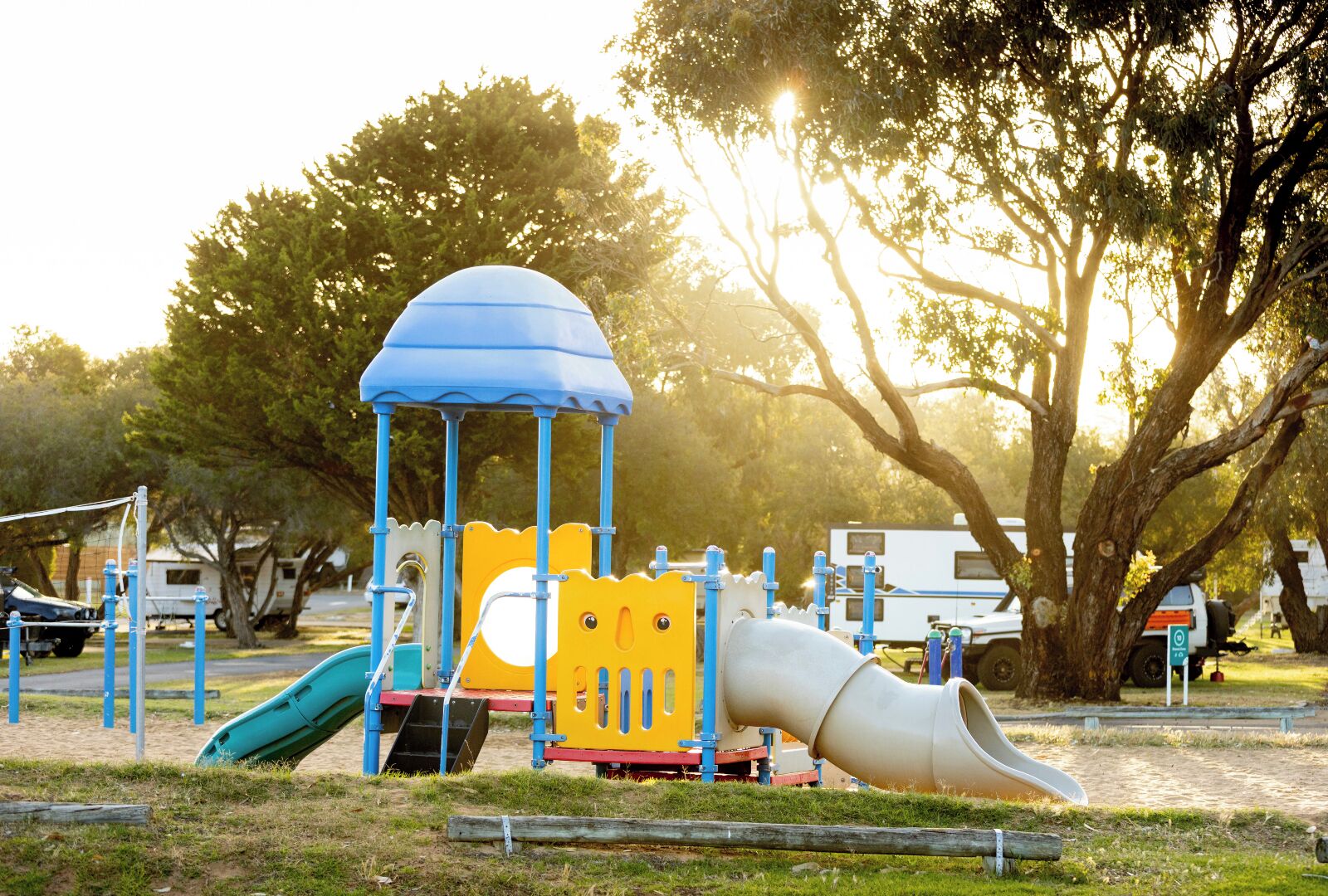 A playground with a slide and climbing structures, surrounded by trees and parked vehicles.