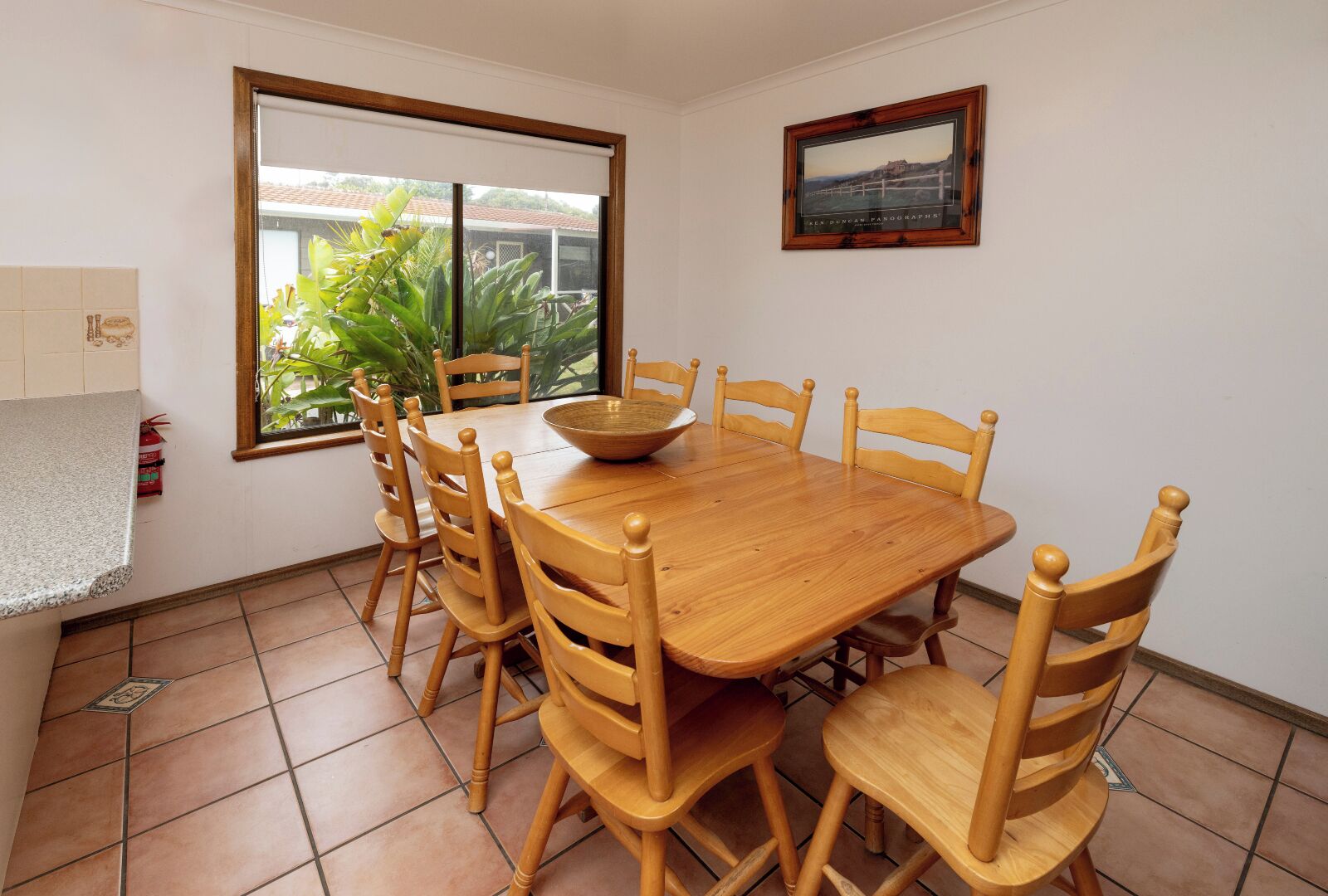 Un comedor con una mesa y sillas de madera, una ventana con vistas a la vegetación y un cuadro en la pared.