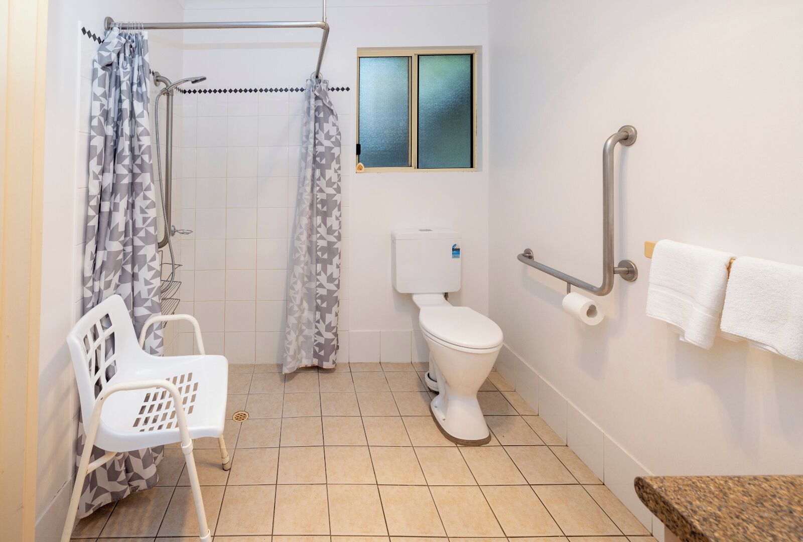 A bathroom with a shower, toilet, grab bar, and a chair.