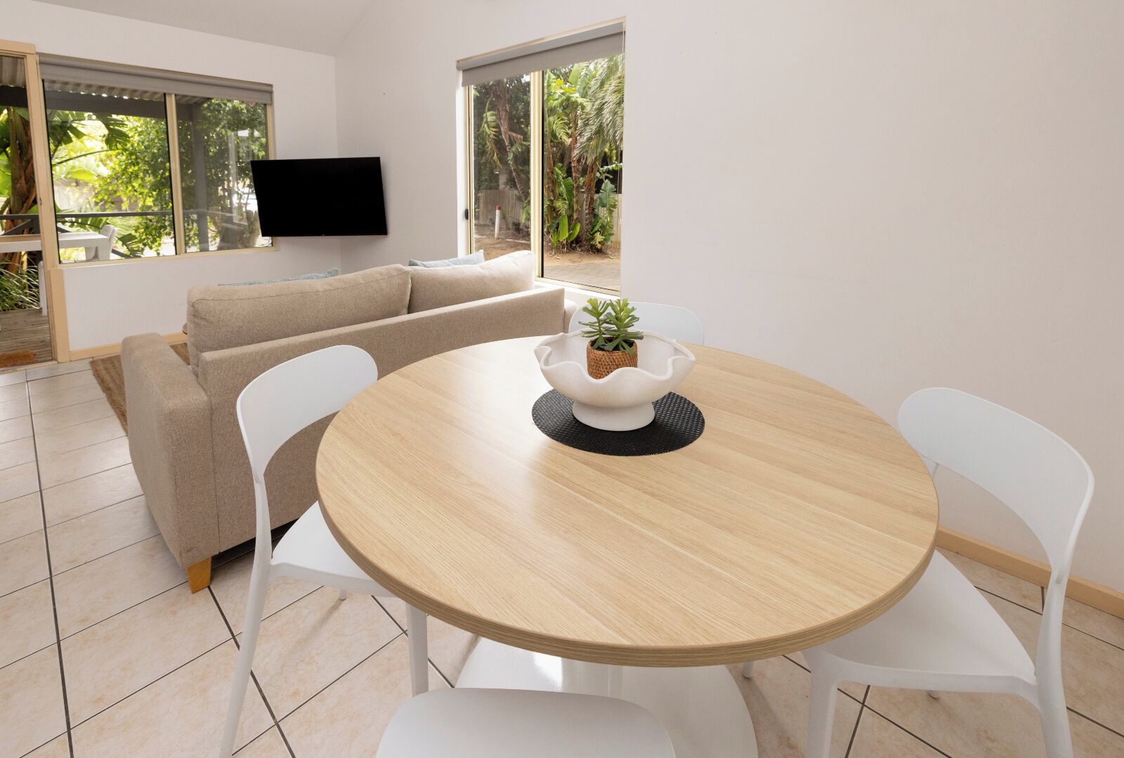 A modern living room with a beige sofa, a round wooden dining table with white chairs, a flat-screen TV mounted on the wall, and large windows with a view of greenery outside.