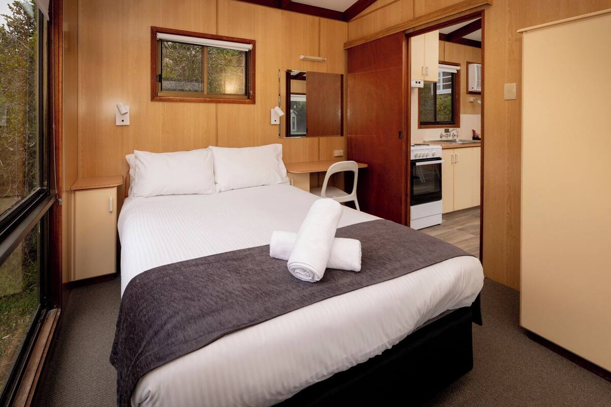 Een net opgemaakt bed met een grijze deken en opgerolde handdoeken, houten wanden, een klein bureau en een kitchenette op de achtergrond.