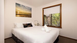 Un lit bien fait, avec du linge de lit blanc, un seul oreiller et une serviette enroulée. Un tableau représentant un coucher de soleil est accroché au mur au-dessus du lit, et une fenêtre offre une vue sur la verdure.