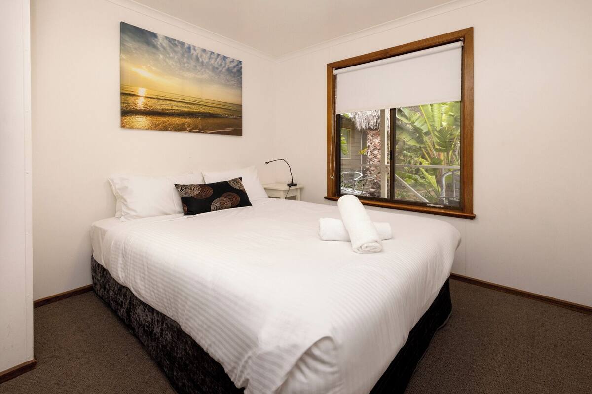 Een keurig opgemaakt bed met witte beddengoed, een enkel kussen en een opgerolde handdoek. Daarboven hangt een schilderij van een zonsondergang en is er een raam met uitzicht op groen.
