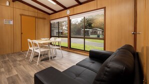 Une pièce avec un canapé en cuir noir, une table à manger avec des chaises blanches et de grandes fenêtres donnant sur une maison et des arbres.