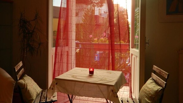 A room with a table covered by a white tablecloth, a red candle on the table, and a view of a balcony with a red railing. - Bed & Roses (Montesilvano)