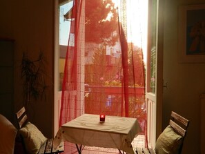 Een kamer met een tafel bedekt met een witte tafelkleed, een rode kaars op de tafel, en uitzicht op een balkon met een rode balustrade.