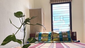 Ein Schlafzimmer mit Bett, Fenster mit Jalousien, Nachttisch mit Büchern und einer Pflanze.