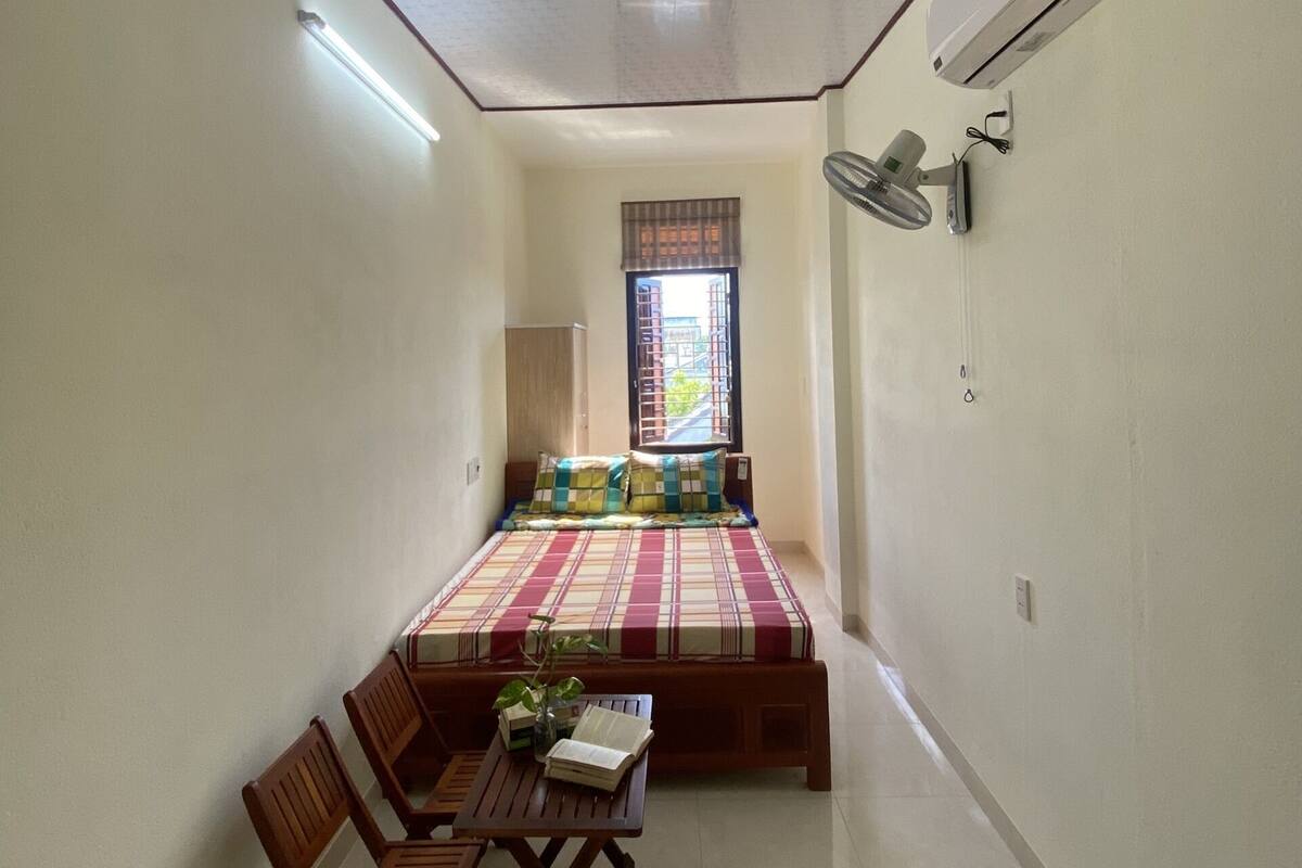 A small, narrow room with a bed, a table with chairs, a window with a view, and an air conditioning unit.