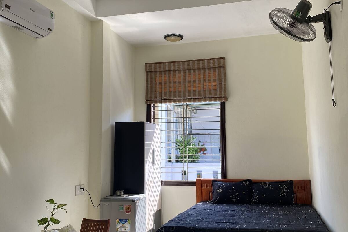 A bedroom with a bed, a desk with a chair, a radiator, a window with blinds, and a wall-mounted fan.