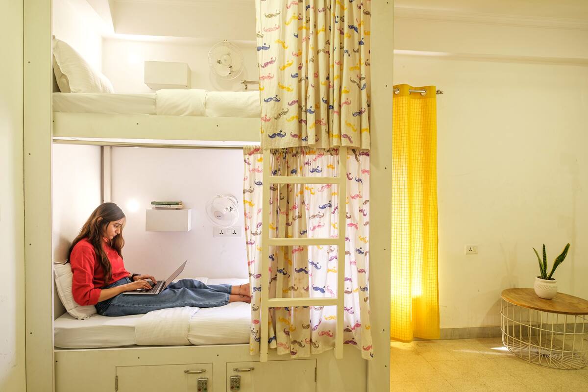 A bunk bed room with a person using a laptop, a small table with a plant, and a yellow curtain.